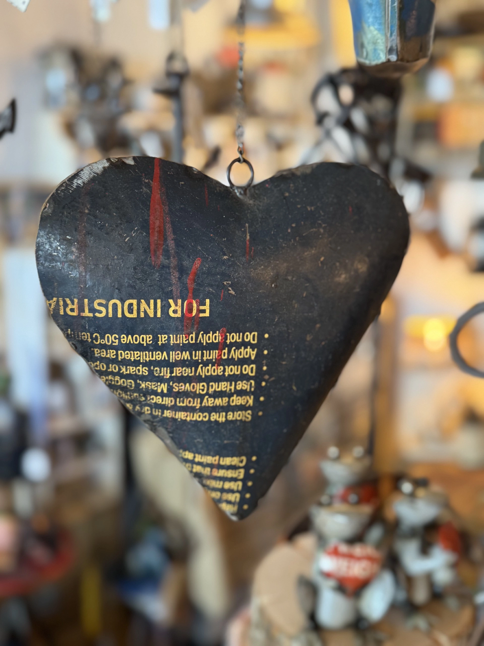 Schwarzes, herzförmiges Metallschild mit gelber und roter Schrift hängt an einem Haken vor unscharfem Hintergrund mit Figuren