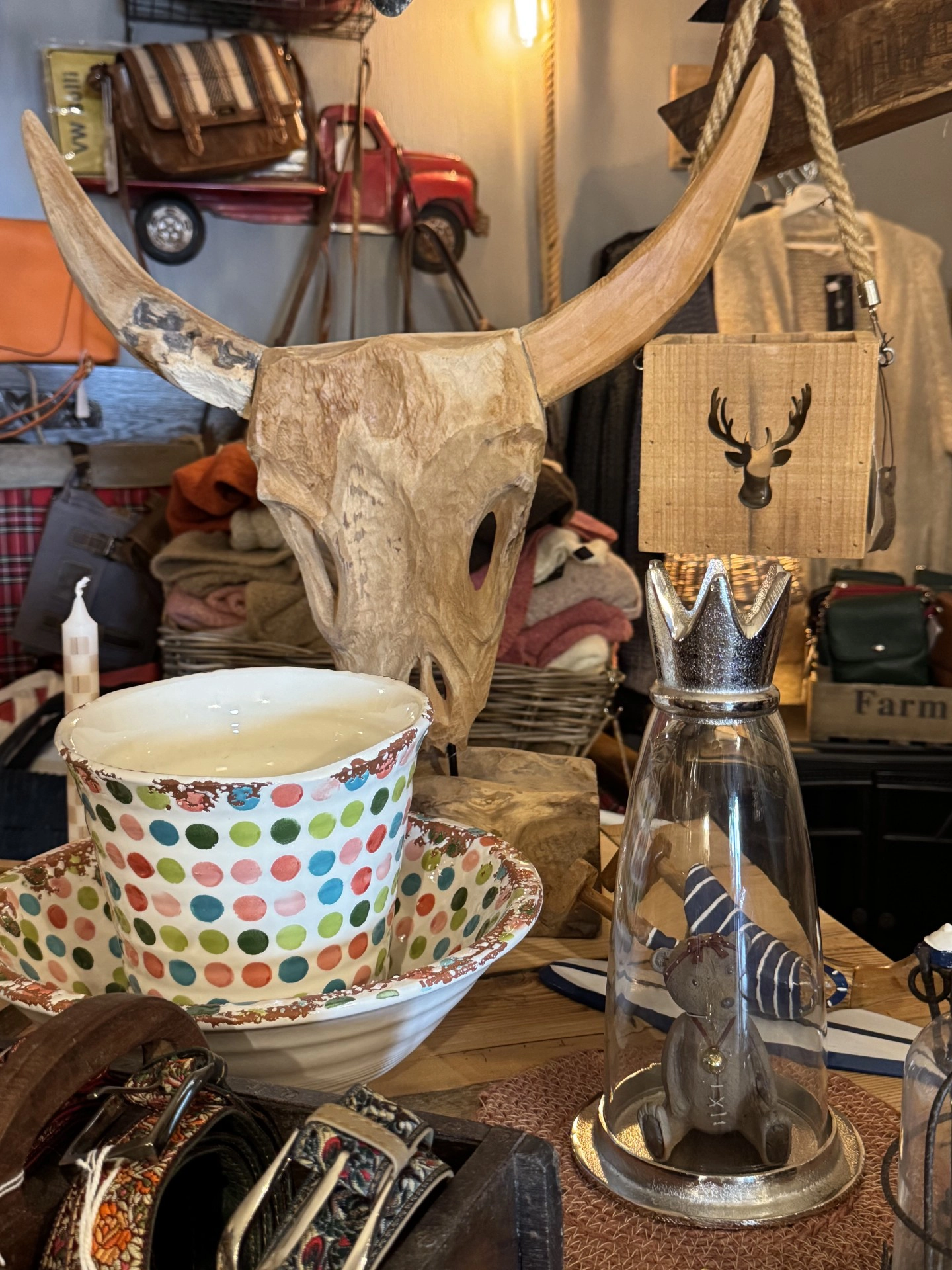 Holzskulptur eines Stierkopfs mit langen Hörnern auf einem Tisch mit buntem Punktmusterbecher und Glasflasche mit Kronendeckel und Stofftier im Inneren
