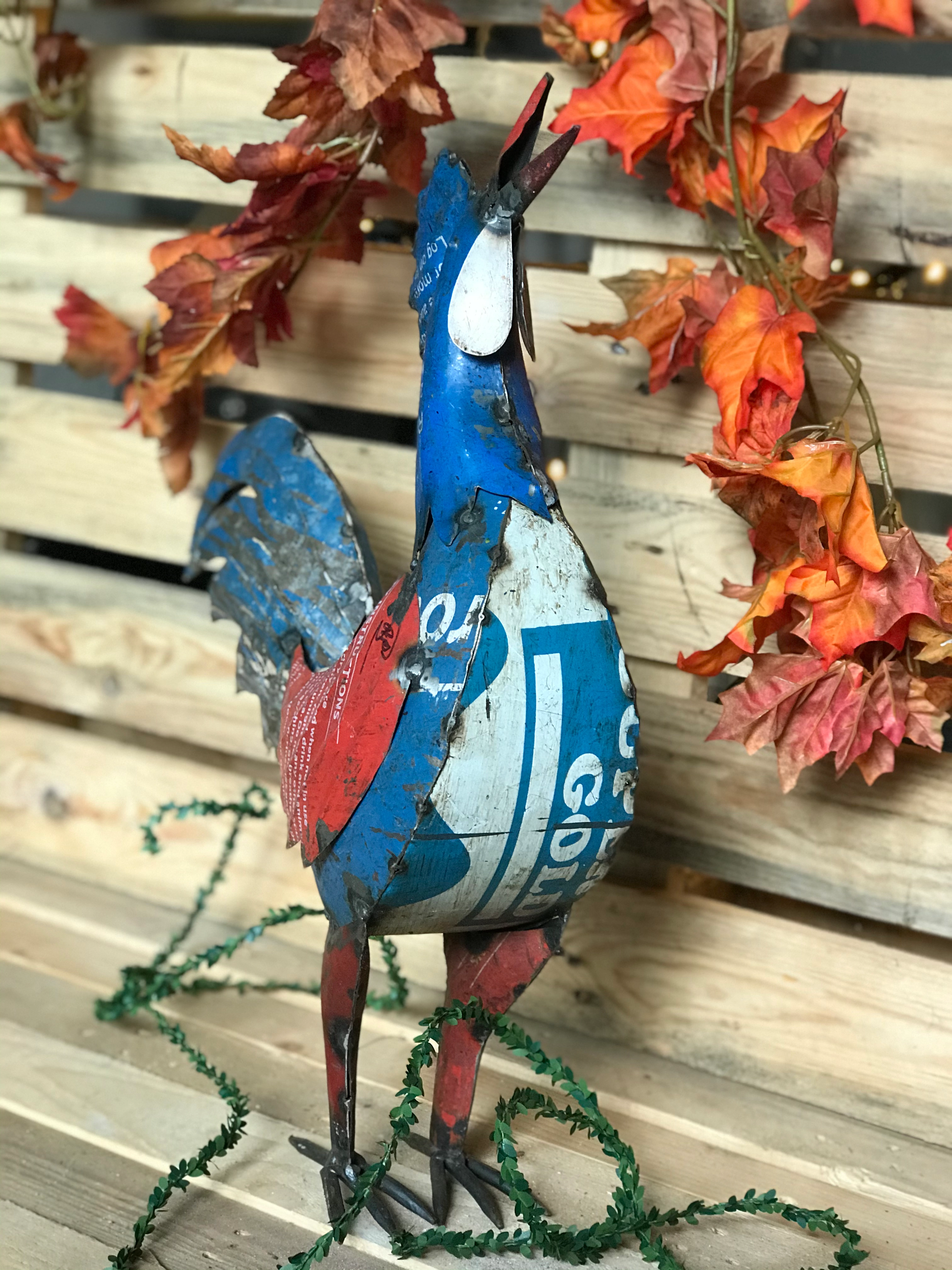 Metallskulptur eines Hahns aus recyceltem, bemaltem Blech vor einer Holzwand mit herbstlichen Blättern