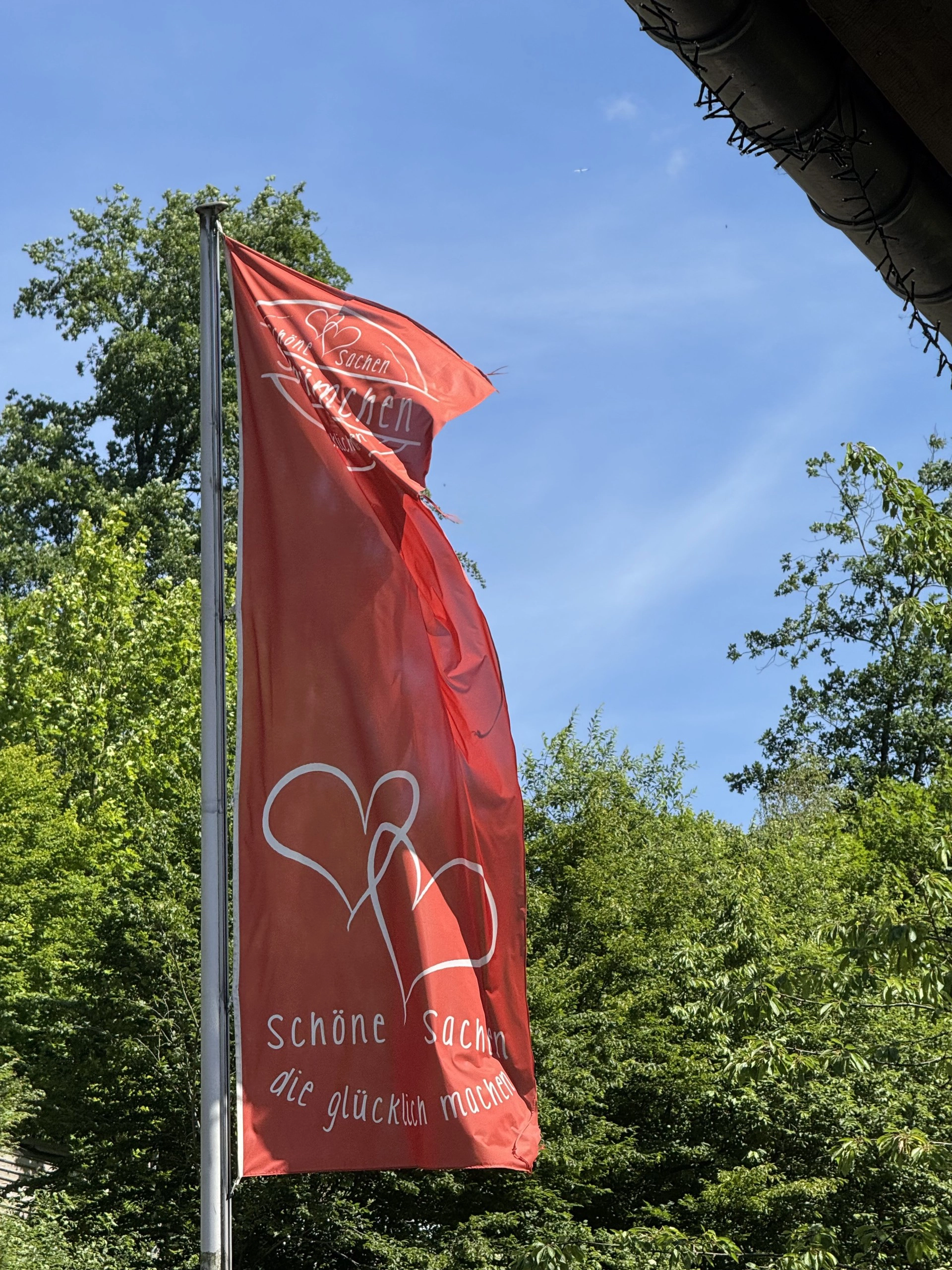 Rote Fahne mit weißen Herzen und der Aufschrift 'schöne Sachen die glücklich machen' vor grünem Laub und blauem Himmel