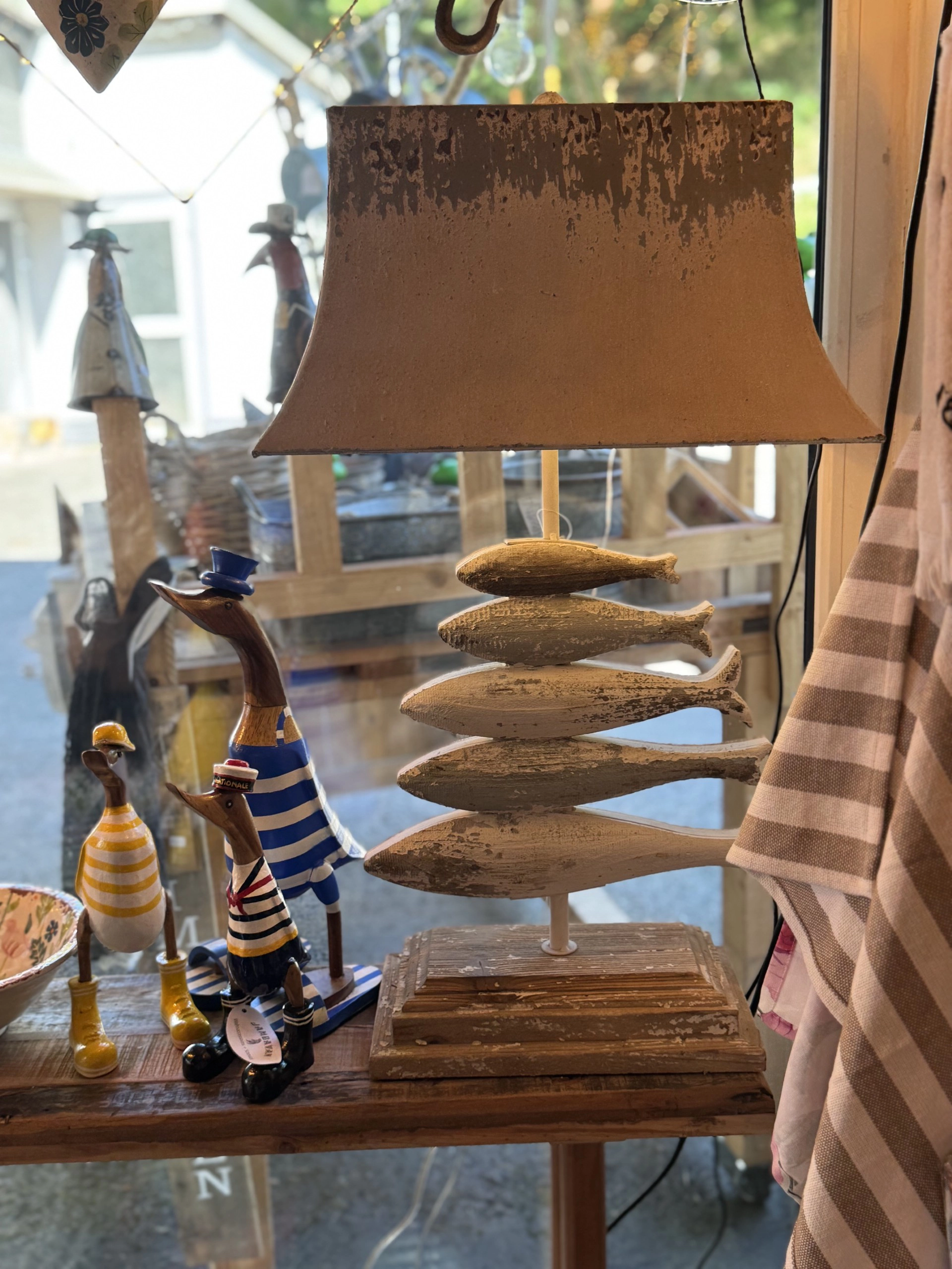 Tischlampe mit Lampenschirm und Sockel aus übereinander gestapelten Holzfischen neben drei hölzernen, bunt bemalten Vogel-Figuren auf einem Fensterbrett