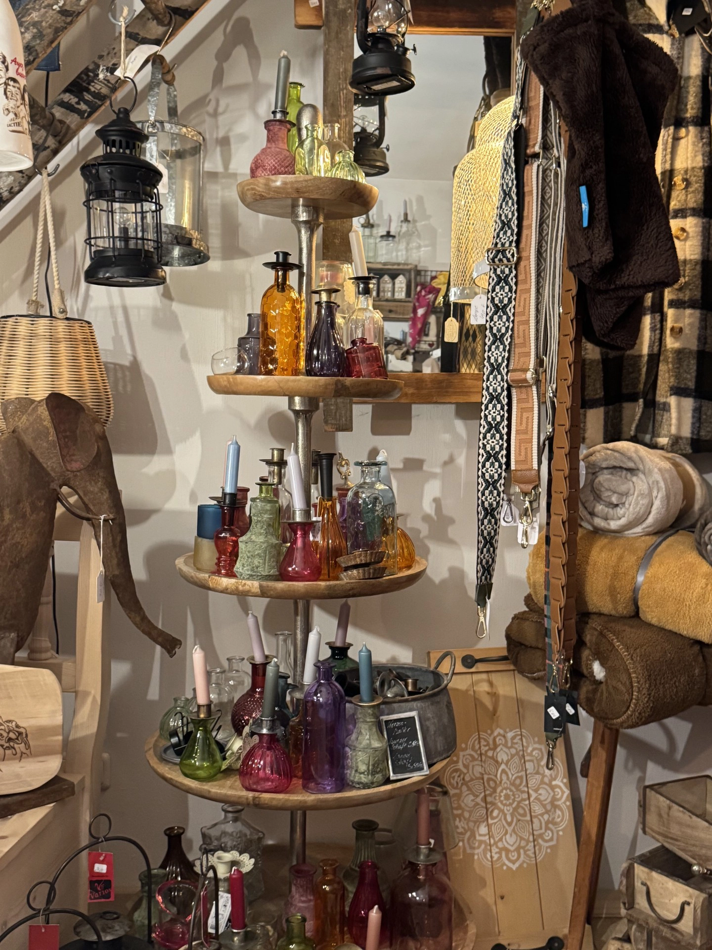 Mehrstöckiges Holzregal mit bunten Glasflaschen und Kerzen in einem Raum mit weiteren Wohnaccessoires und Textilien