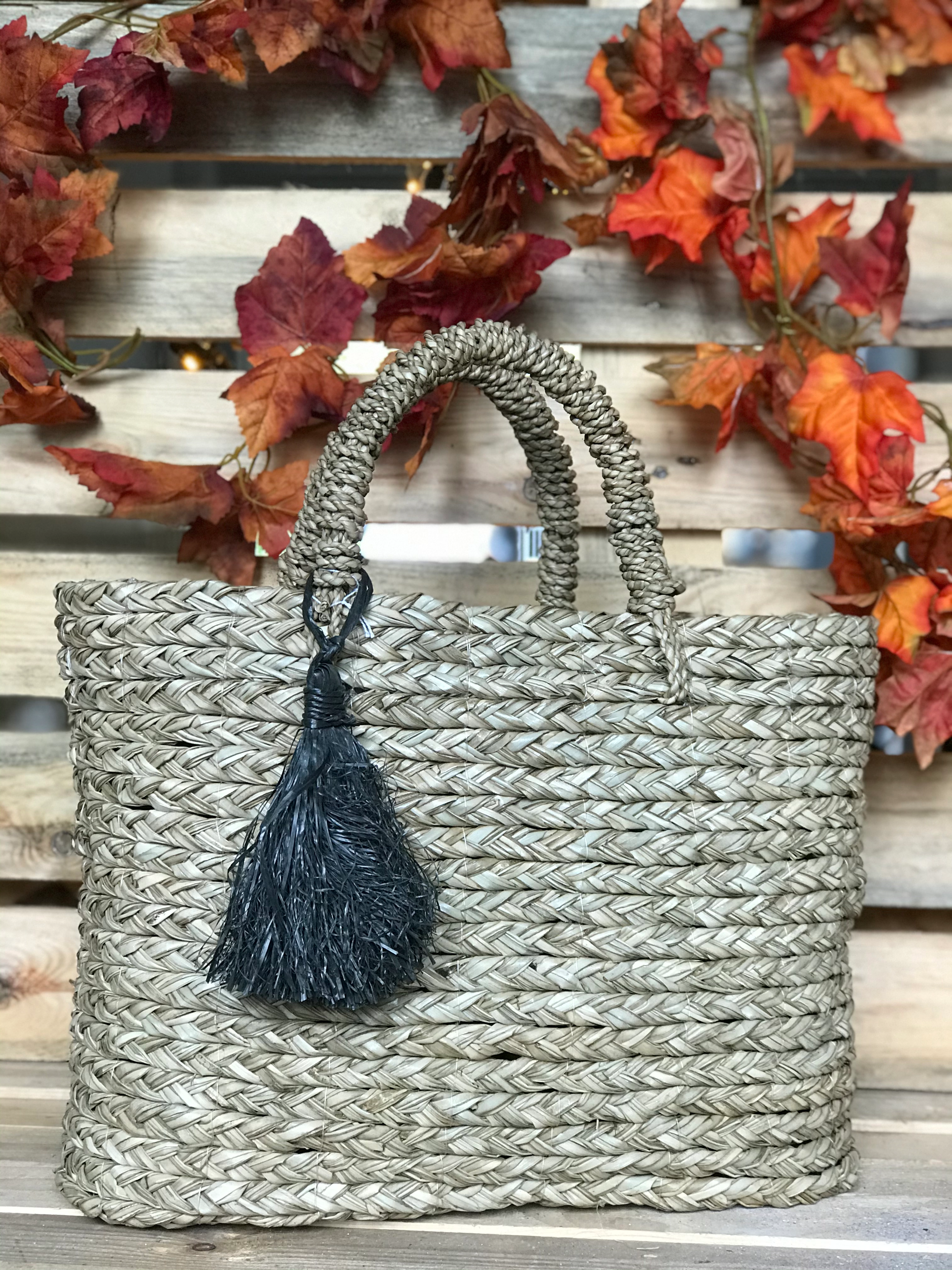 Geflochtene Tasche mit zwei Henkeln und einem schwarzen Quastenanhänger vor einer Holzpalette mit herbstlichen Blättern